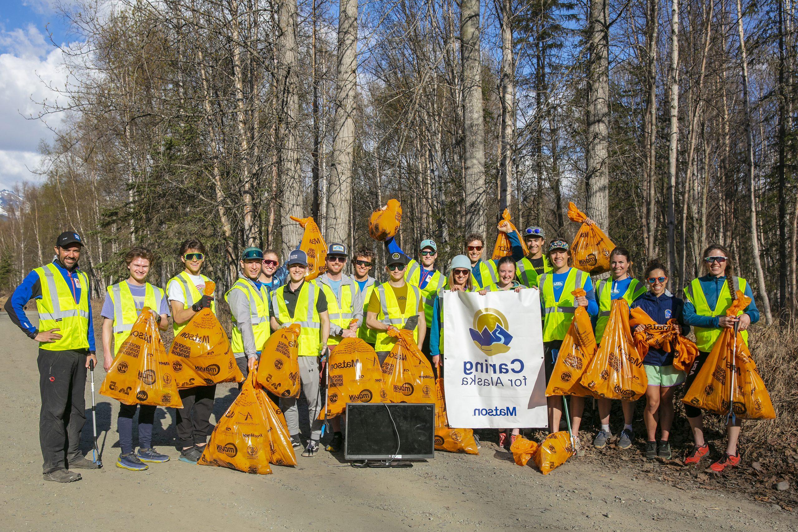 Volunteers wearing yellow safety vests pose for a group picture with their full orange trash bags and 关爱阿拉斯加 banner.